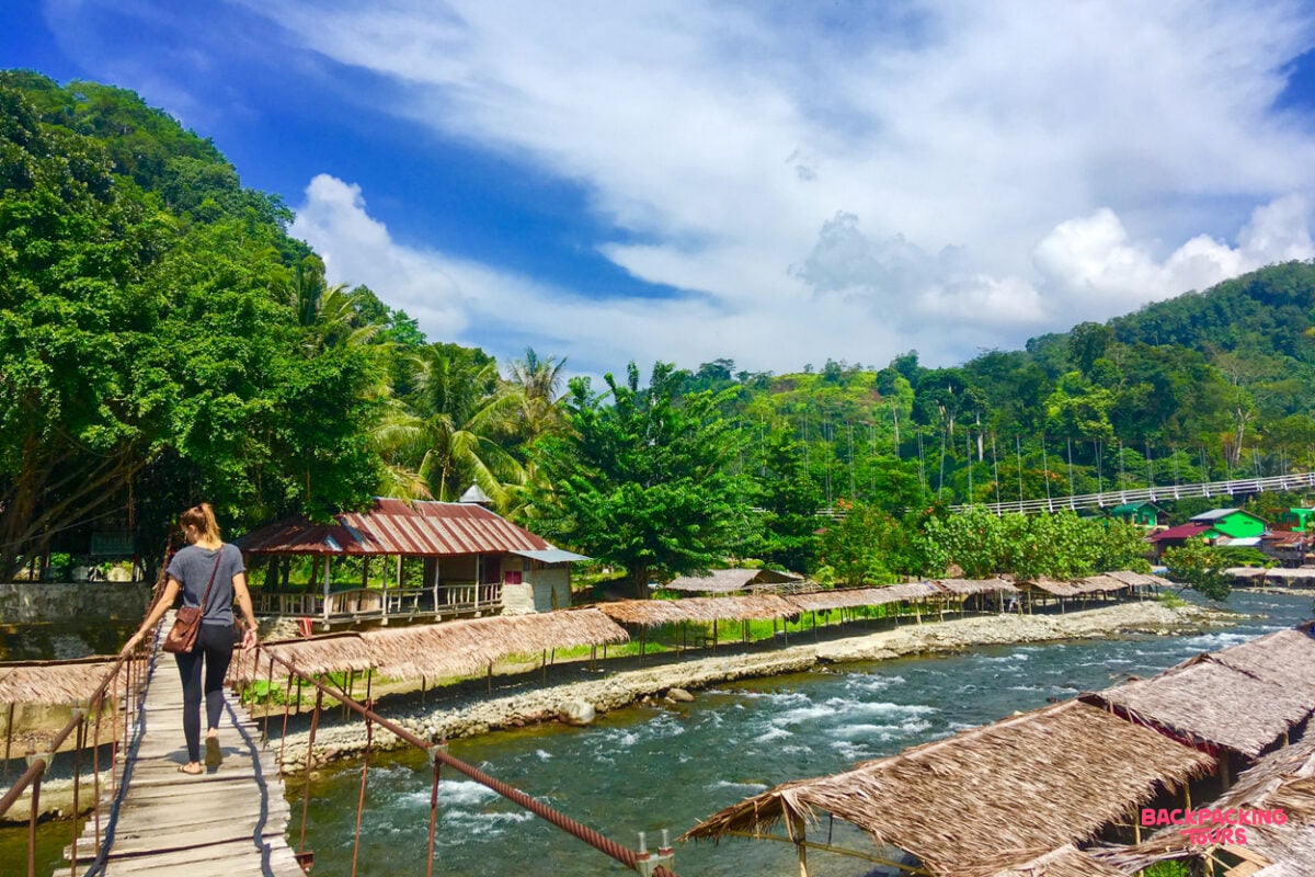 A backpacker crosses a wooden bridge over a river, surrounded by lush jungle in Bukit Lawang.