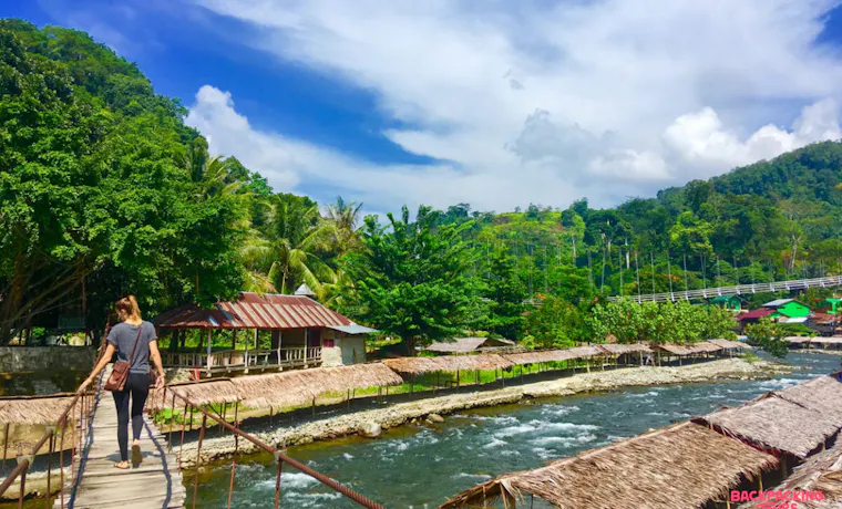 A backpacker crosses a wooden bridge over a river, surrounded by lush jungle in Bukit Lawang.