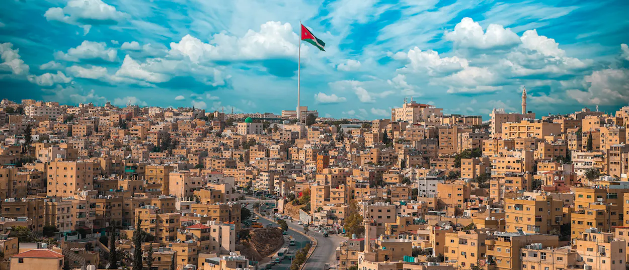 Vast Amman cityscape with numerous buildings, a central road, and the Jordanian flag against a cloudy sky.