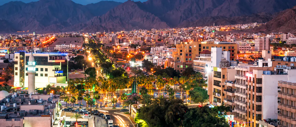 A vibrant city at night in Aqaba, Jordan, with illuminated buildings and streets backed by dark mountains.