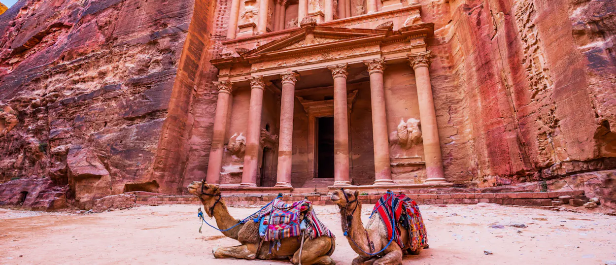 Two colorful saddled camels rest before the magnificent, ancient Treasury carved into Petra's red cliffs.
