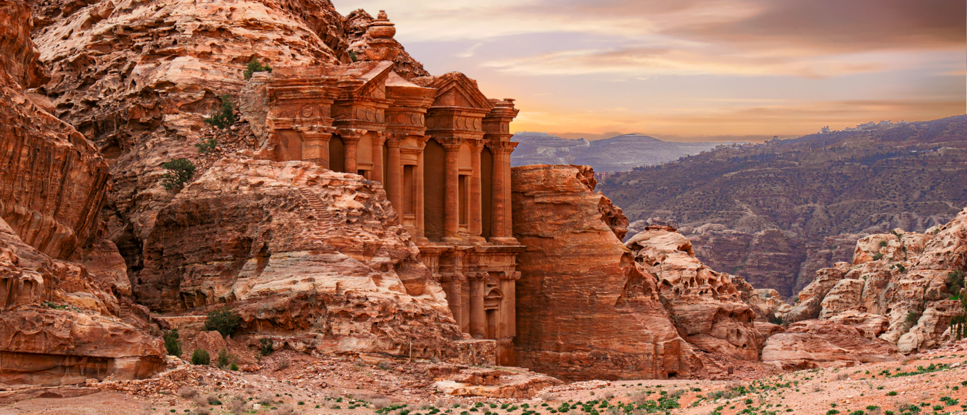 The ancient Monastery at Petra carved into a towering red rock cliff at sunset.