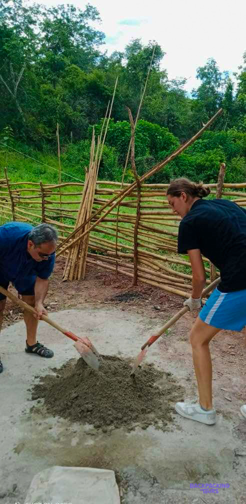 Two volunteers turn a heap of cement with shovels beside a new bamboo fence.
