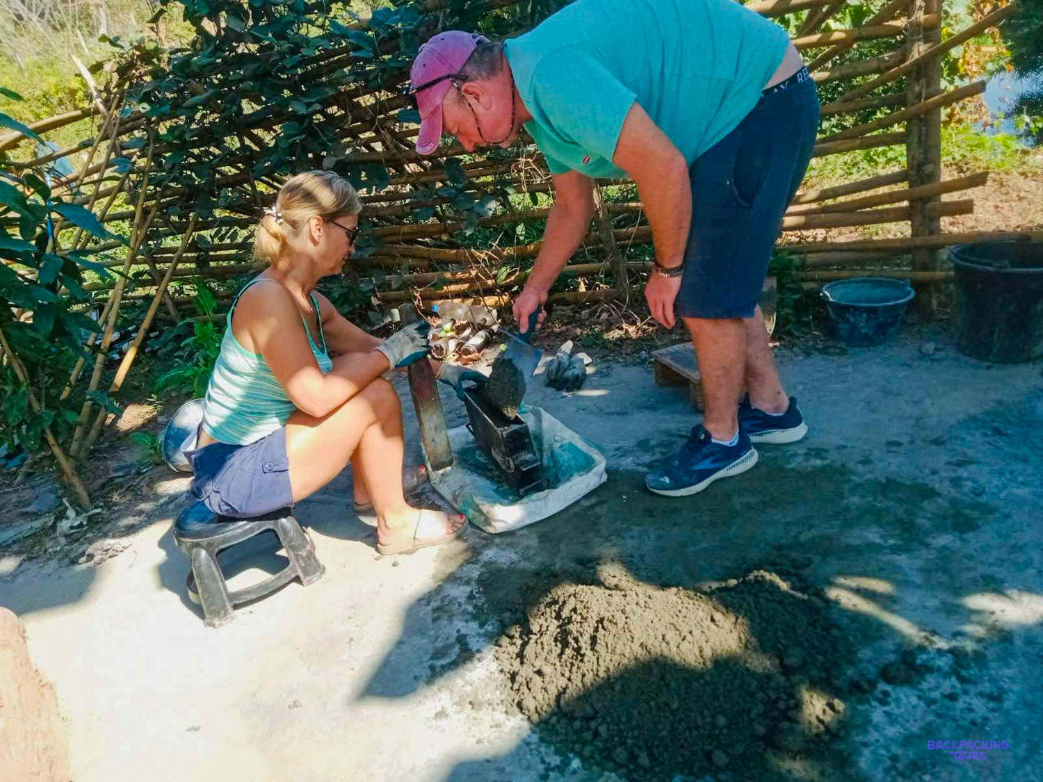 A volunteer in a cap fills a block mould while another holds it steady on the concrete.