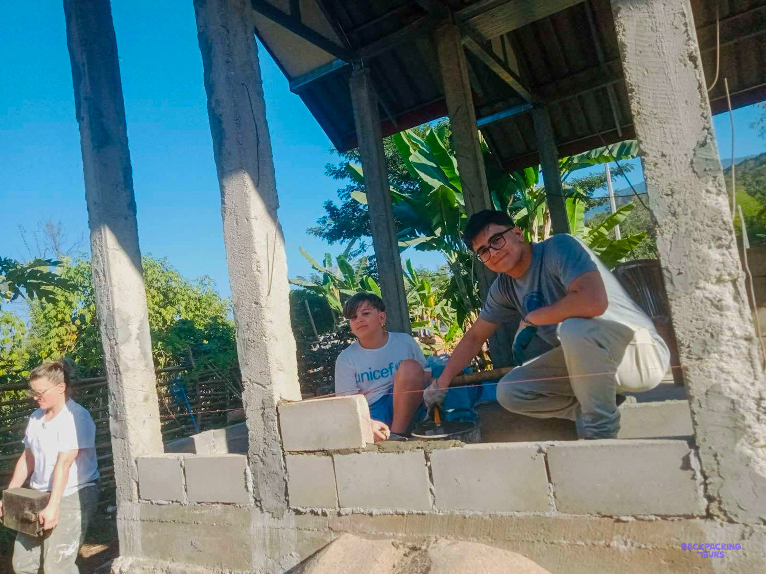 Young volunteers lay cement blocks between concrete pillars to build a new wall.