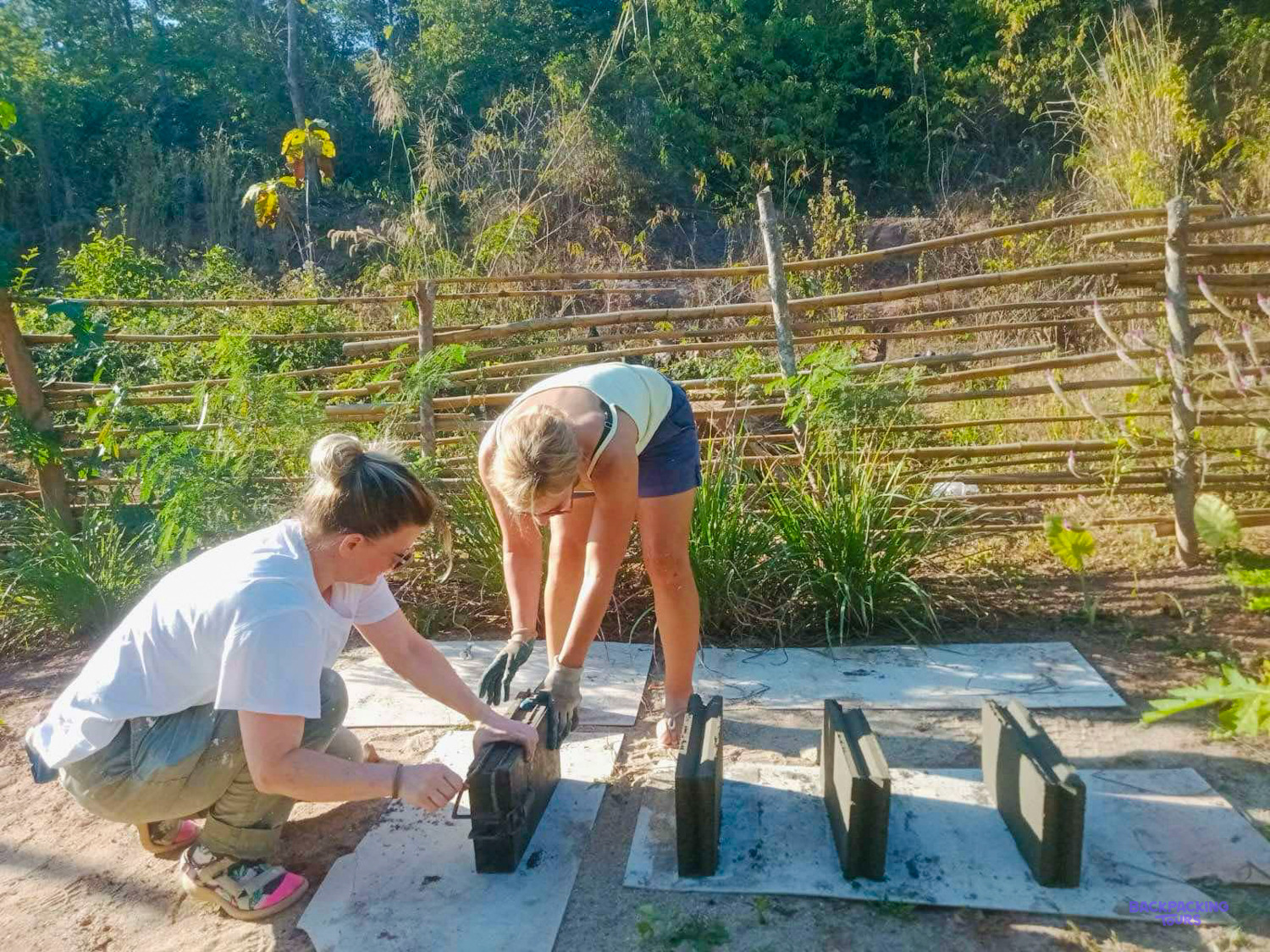 Two volunteers in gloves lift fresh cement blocks out of their moulds beside a bamboo fence.
