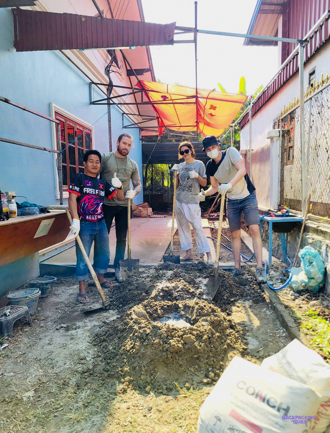 Volunteers in gloves turn a pile of cement with shovels in a narrow alley between houses.
