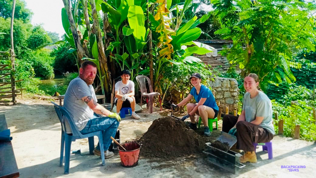 Volunteers on plastic stools mix cement and fill block moulds in a leafy village yard.