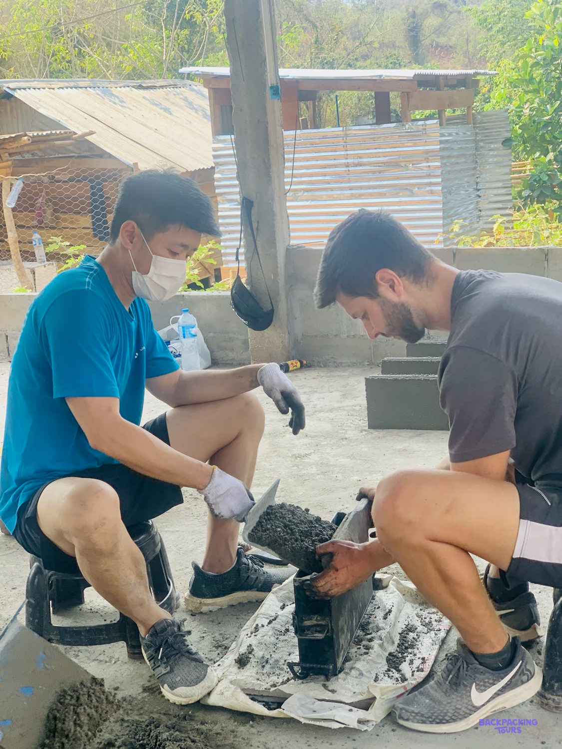 Two volunteers in masks and gloves press cement into a block mould on a concrete floor.