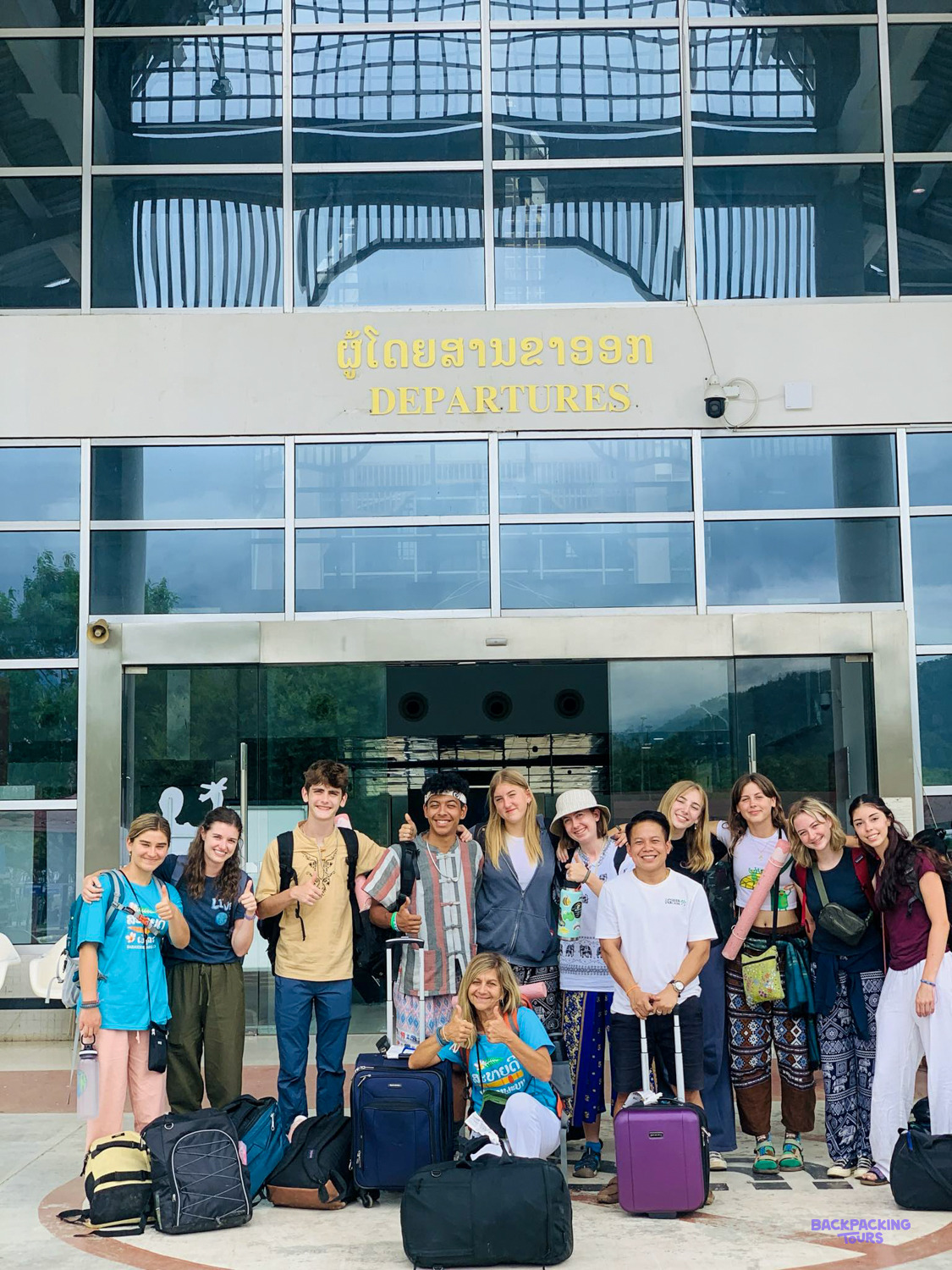 A smiling group of volunteers with their suitcases outside the airport terminal in Laos.