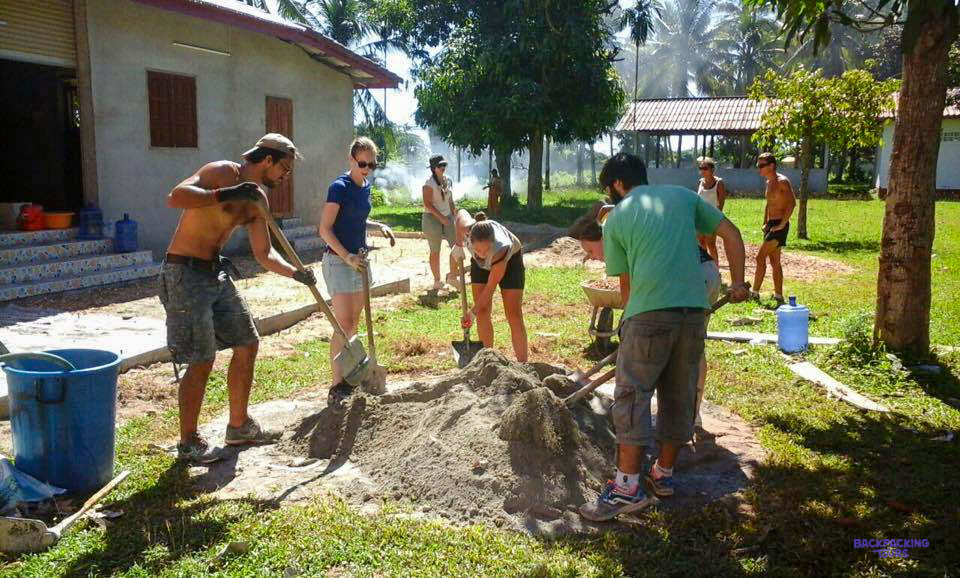 Volunteers shovel sand and gravel on a sunny lawn beside a village house.