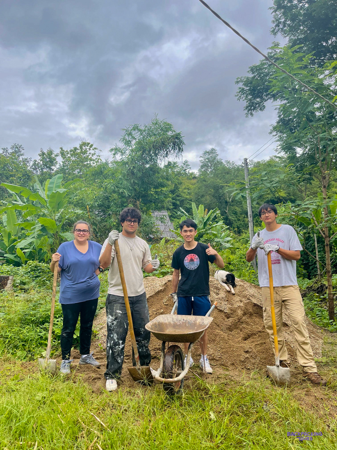 Four volunteers with shovels and a wheelbarrow give a thumbs up beside a pile of sand.