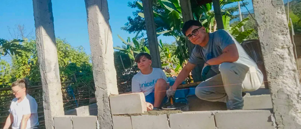 Young volunteers lay cement blocks between concrete pillars to build a new wall.