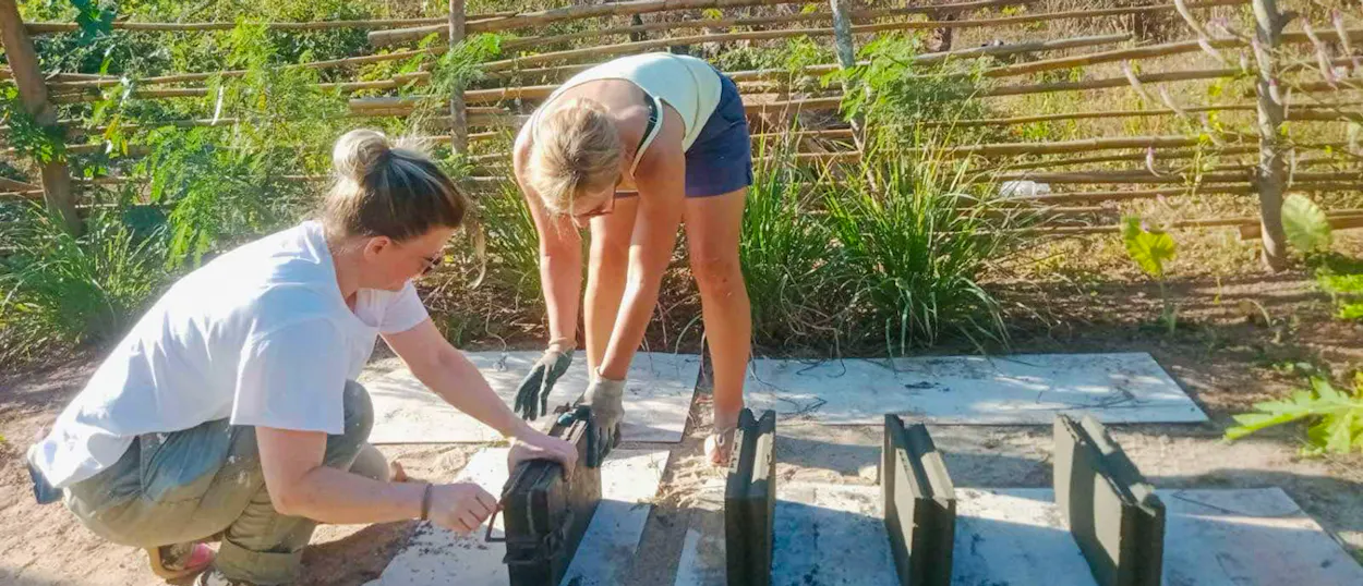 Two volunteers in gloves lift fresh cement blocks out of their moulds beside a bamboo fence.
