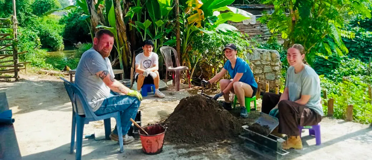 Volunteers on plastic stools mix cement and fill block moulds in a leafy village yard.
