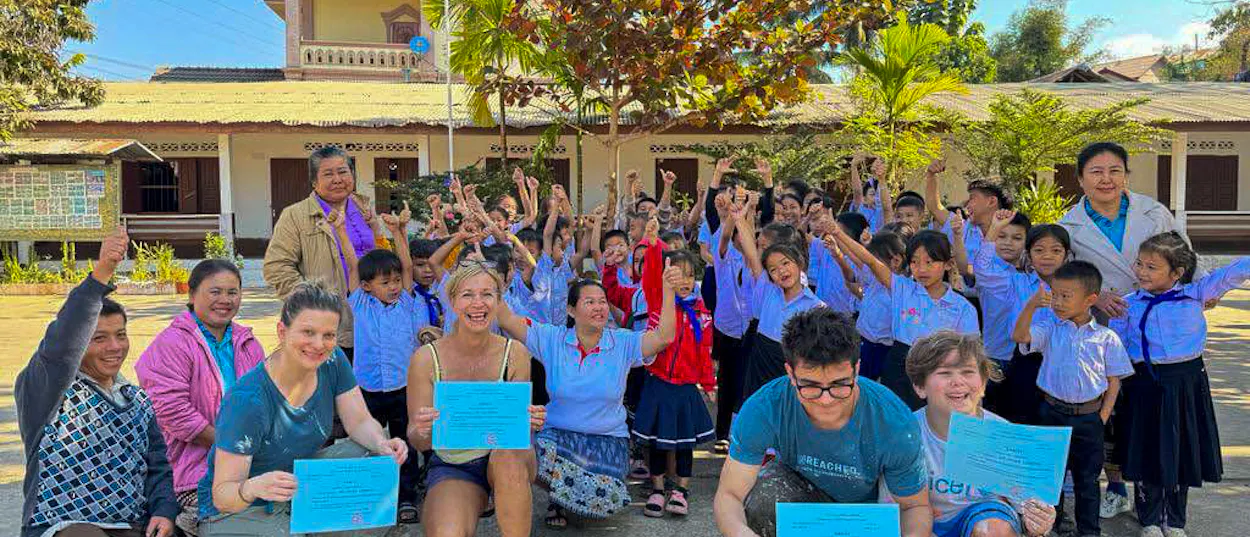 Volunteers hold certificates as cheering schoolchildren and teachers gather outside a Lao school.