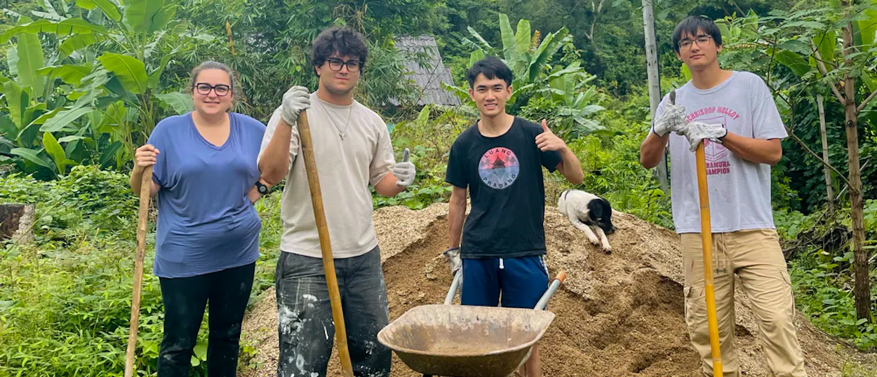 Four volunteers with shovels and a wheelbarrow give a thumbs up beside a pile of sand.