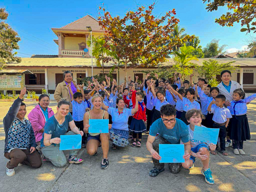 Volunteers hold certificates as cheering schoolchildren and teachers gather outside a Lao school.