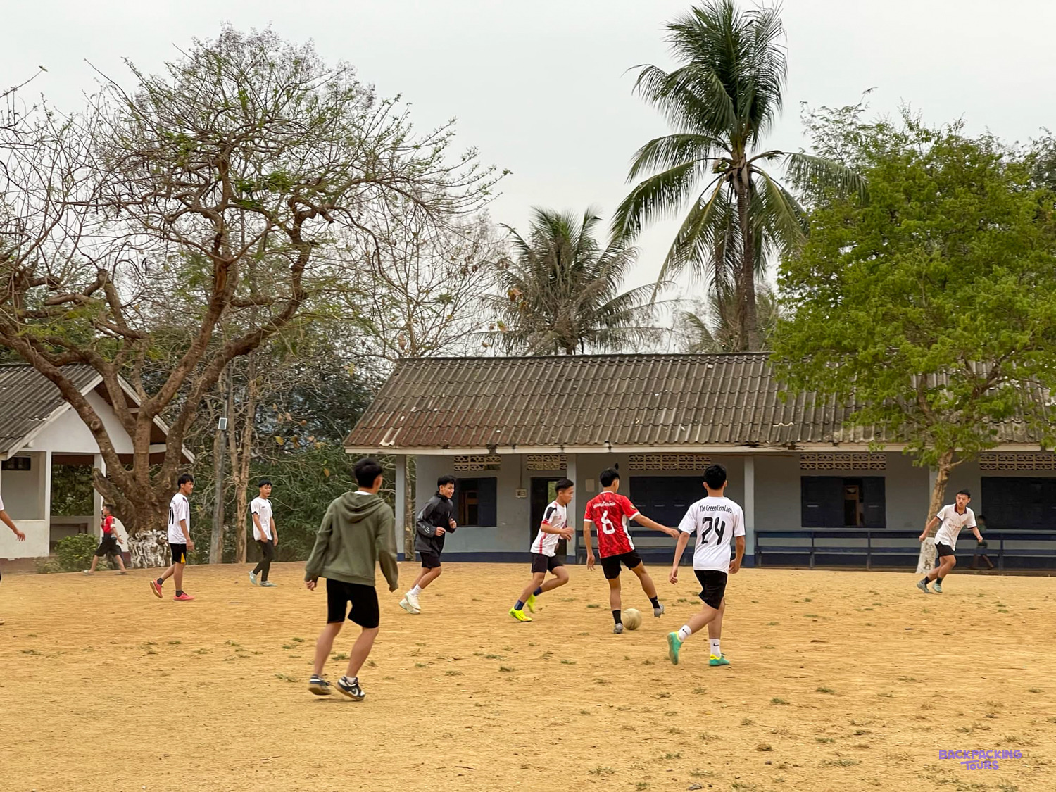 Teenagers play football on a sandy school field in front of a long single-storey classroom block.