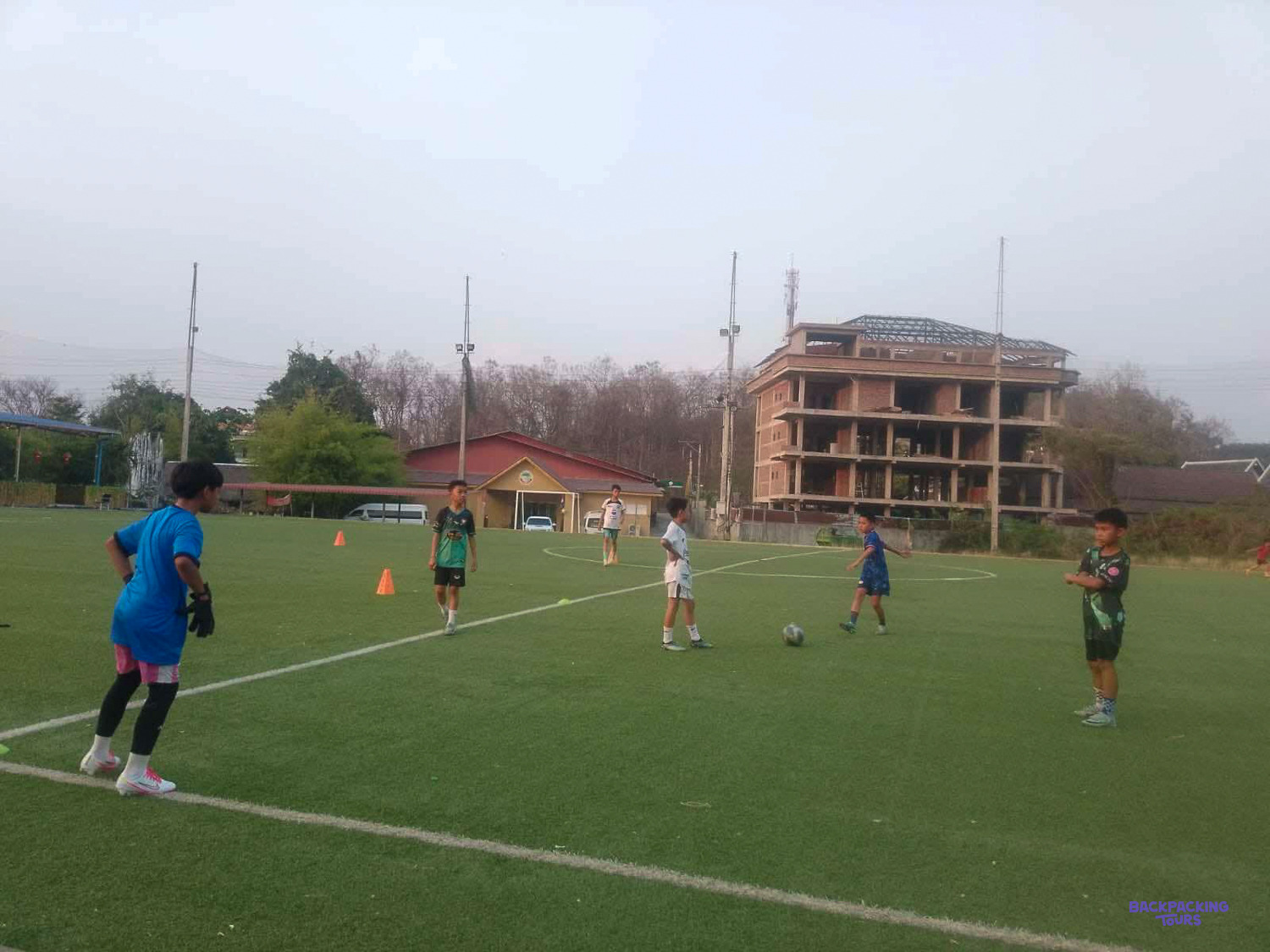 A young goalkeeper in blue waits as players run a drill between orange cones on the pitch.