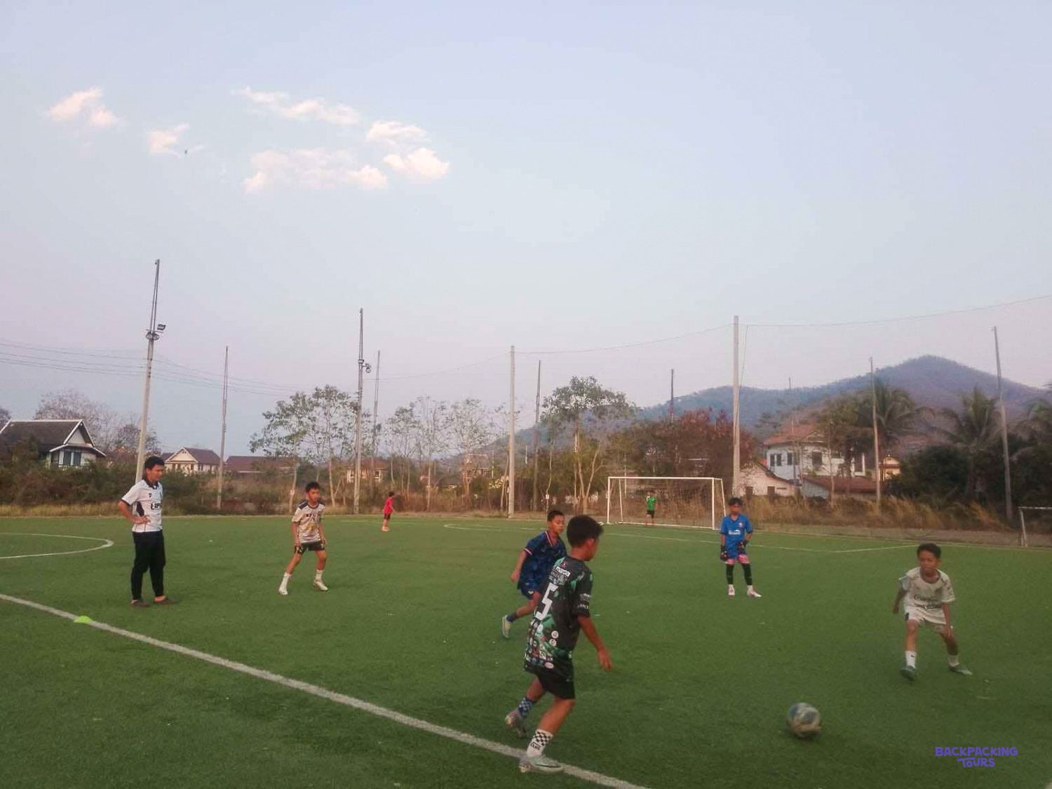 Children play a match on an artificial pitch with green hills rising behind the goal.