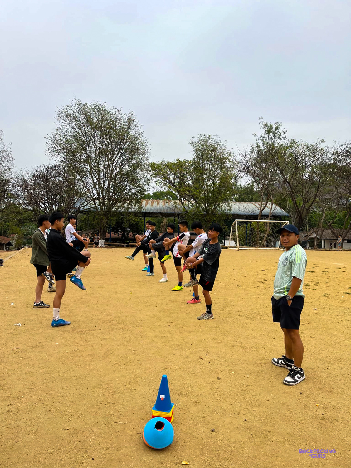 A coach in a cap watches young players stretch and warm up beside a training cone.