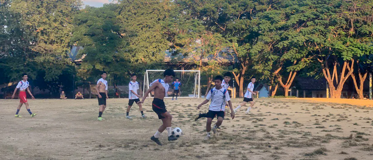 A young player dribbles past a defender on a dirt pitch shaded by tall trees.