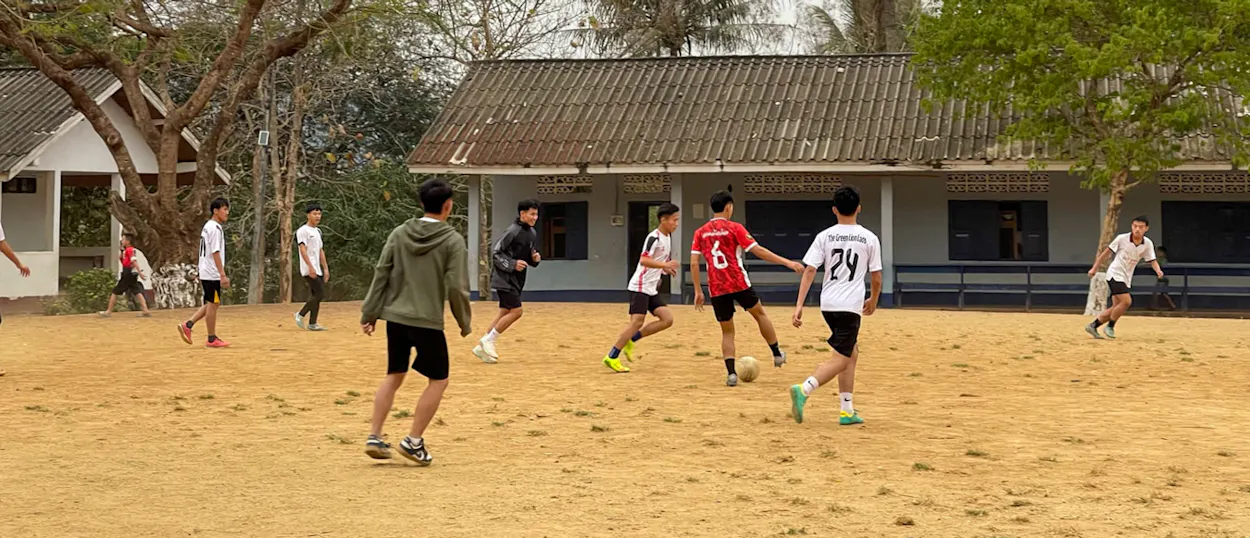 Teenagers play football on a sandy school field in front of a long single-storey classroom block.