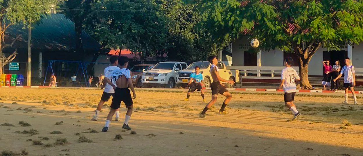 Boys chase a football across a dusty school pitch in the golden evening light in Luang Prabang.
