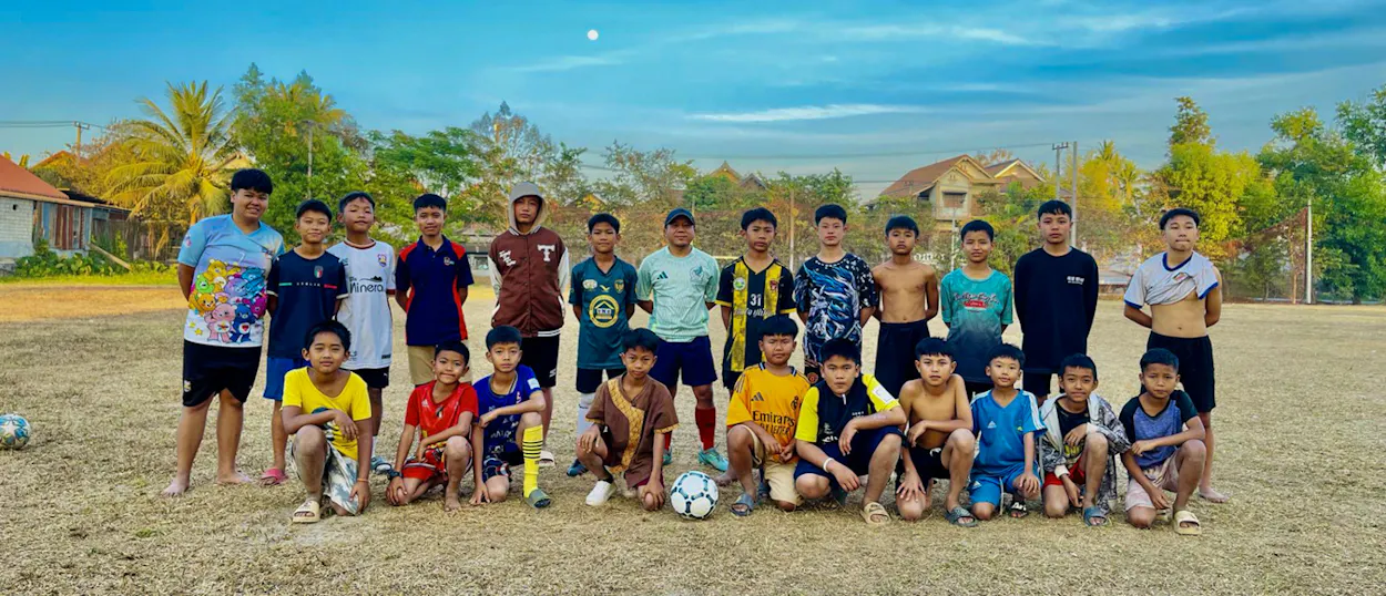 Lao boys of all ages line up with their footballs for a team photo on a sunny pitch.