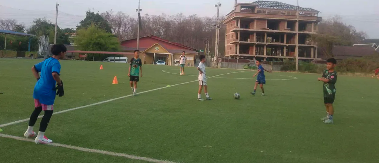 A young goalkeeper in blue waits as players run a drill between orange cones on the pitch.