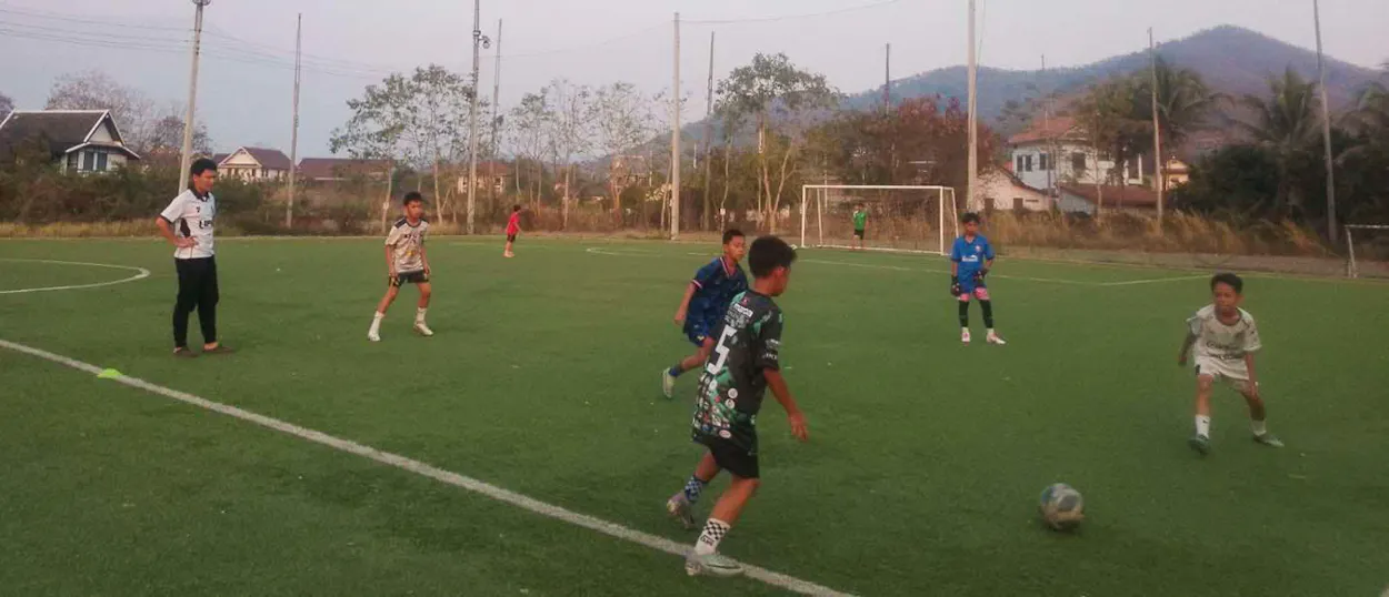 Children play a match on an artificial pitch with green hills rising behind the goal.