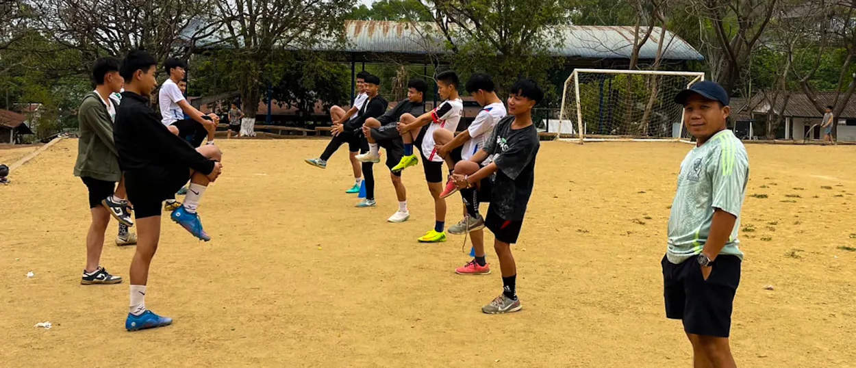 A coach in a cap watches young players stretch and warm up beside a training cone.