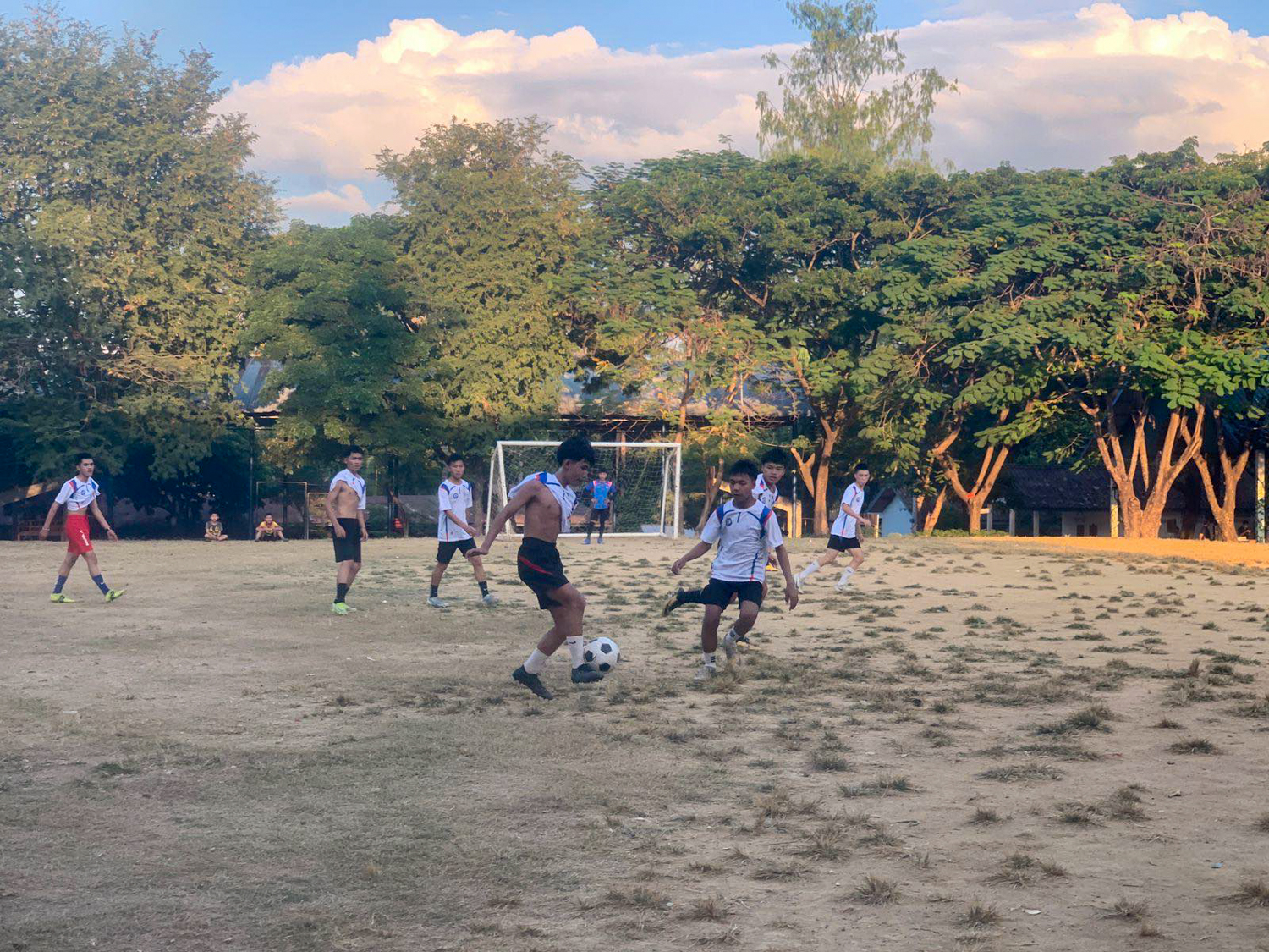 A young player dribbles past a defender on a dirt pitch shaded by tall trees.