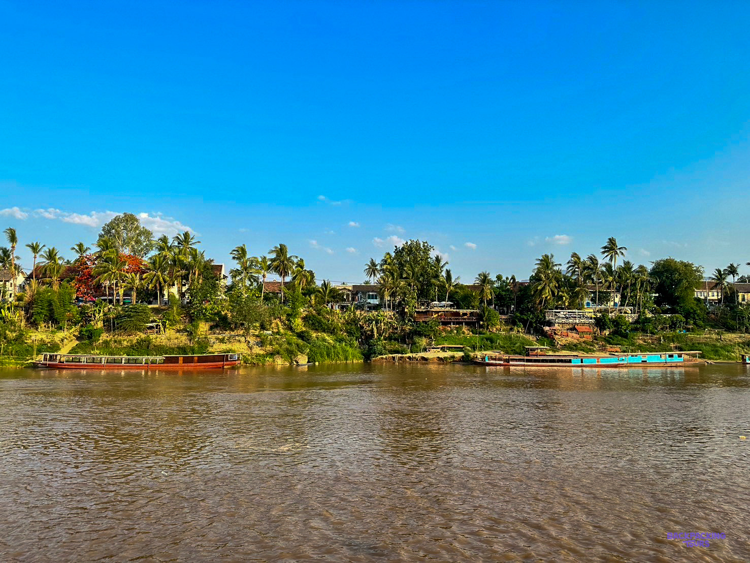 Long wooden boats moored along the tree-lined riverbank at Luang Prabang under a clear blue sky.