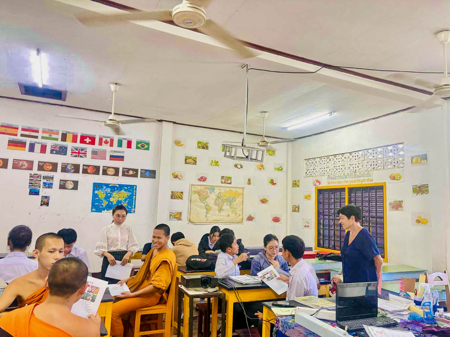 A volunteer stands at the front of a busy class of monks and students under rows of flags.