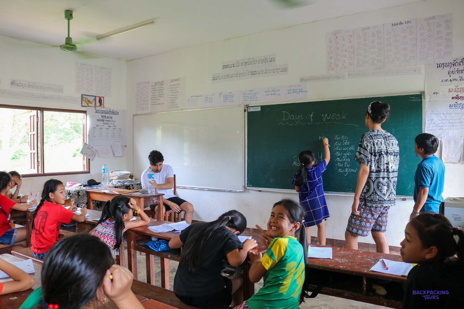 A young pupil writes on a blackboard headed Days of week, helped by a volunteer.