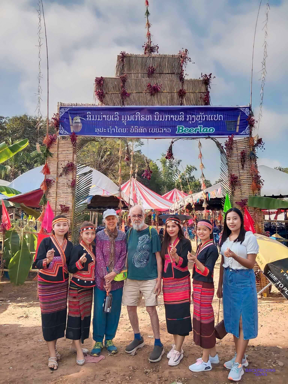 Two volunteers pose with young Lao women in embroidered traditional dress under a festival gateway.