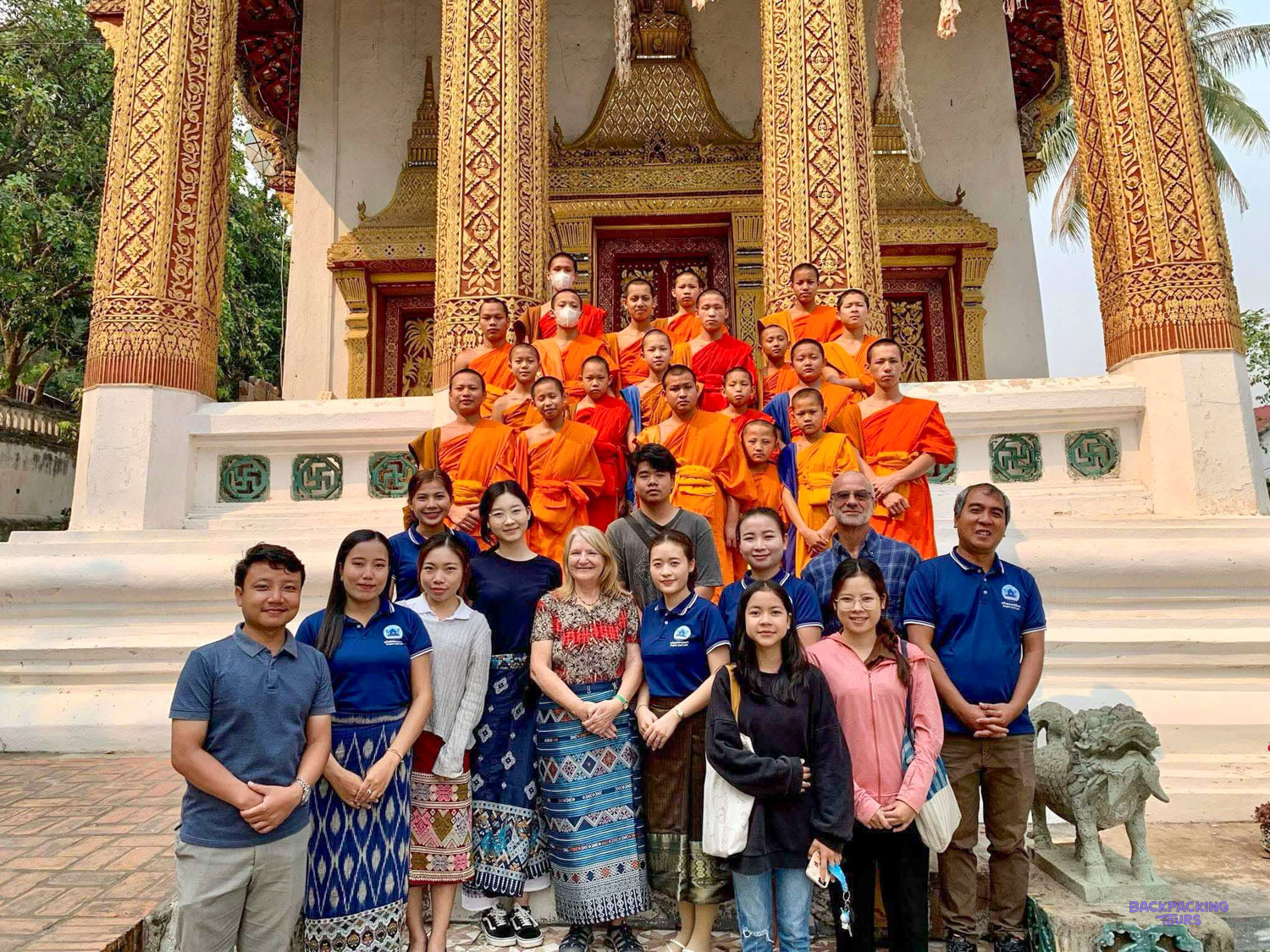 Volunteers and monks in orange robes gather on the steps of an ornate gilded temple.