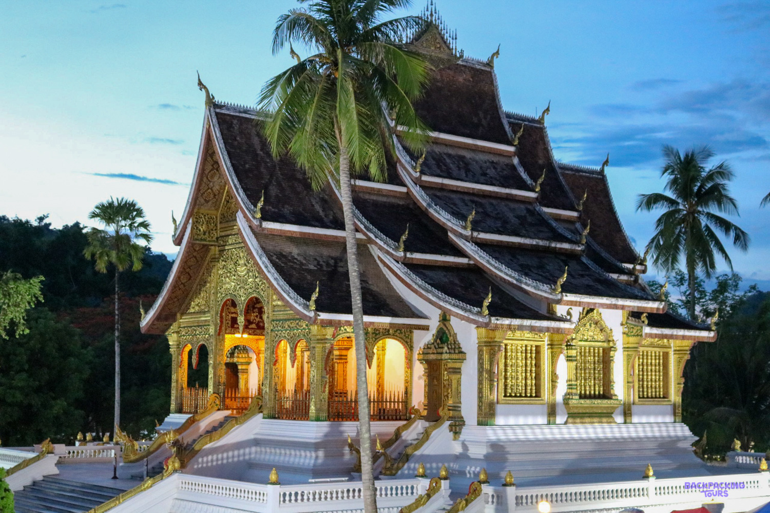 Haw Pha Bang, the gilded royal temple in Luang Prabang, lit up at dusk among palm trees.