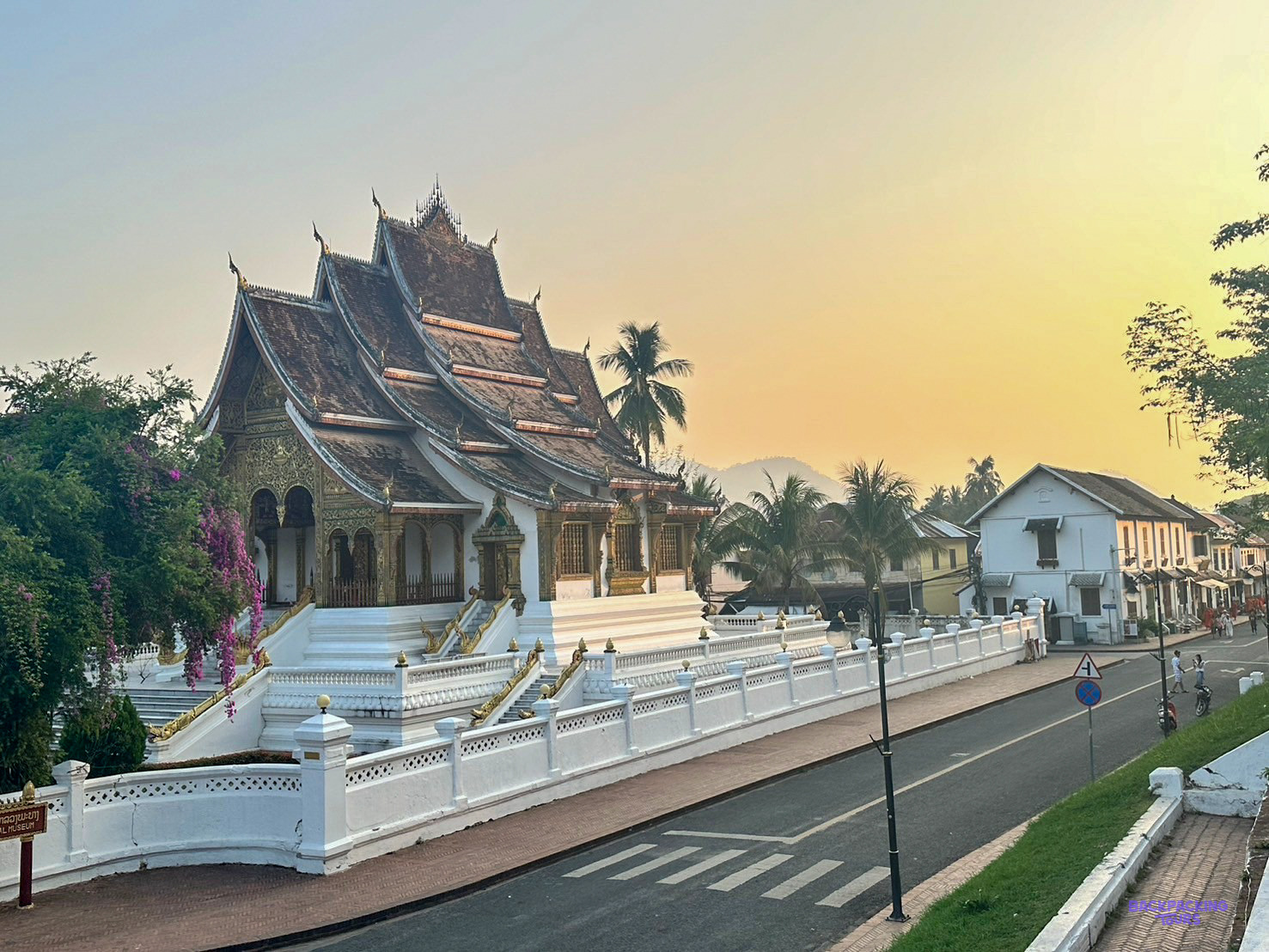 Haw Pha Bang rises above a white wall beside the main street of Luang Prabang at sunset.
