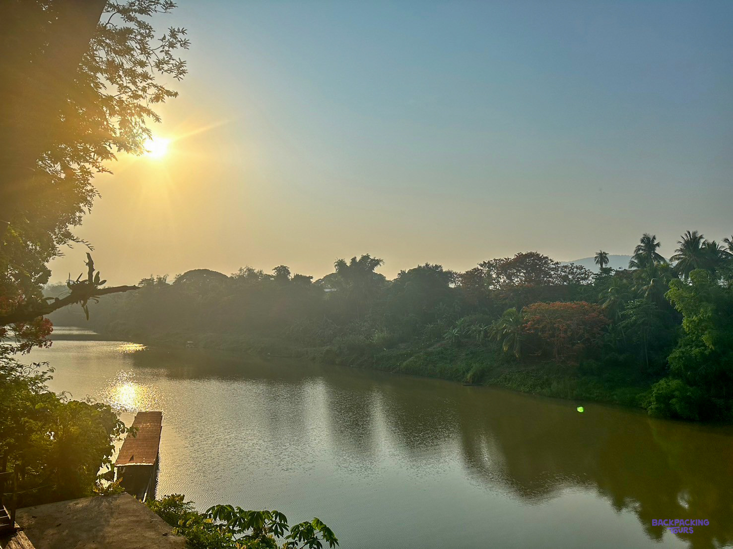 The low sun glows through the haze over a calm river lined with palm trees.