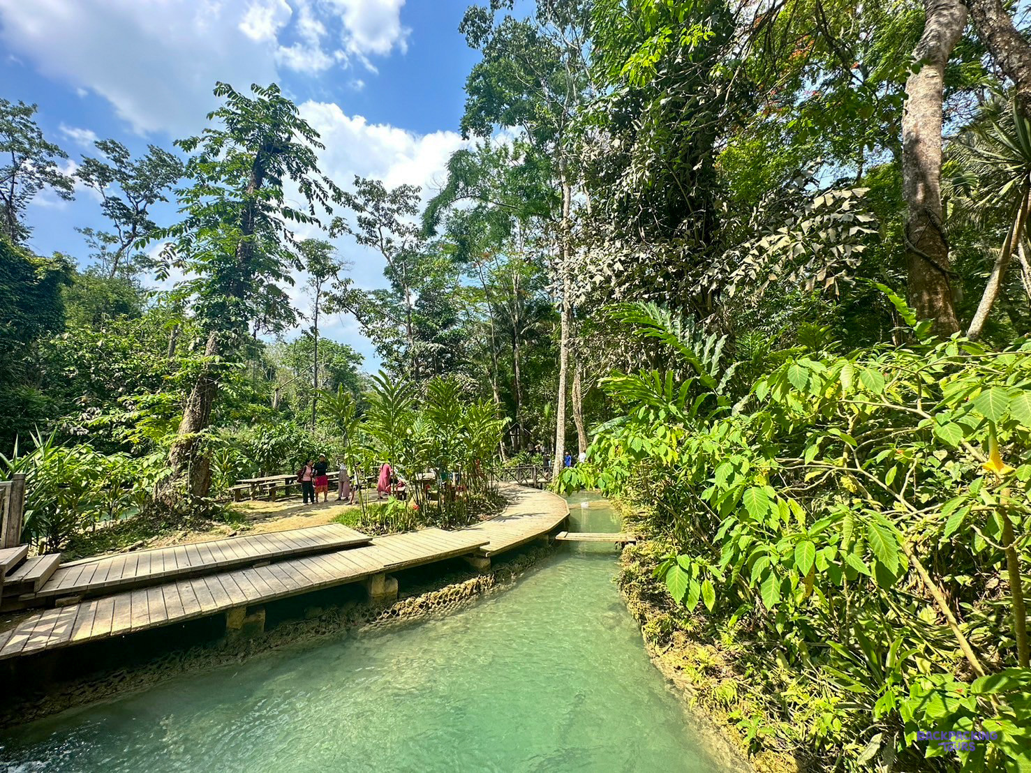 A wooden boardwalk follows a turquoise stream through the forest at the Kuang Si waterfalls.