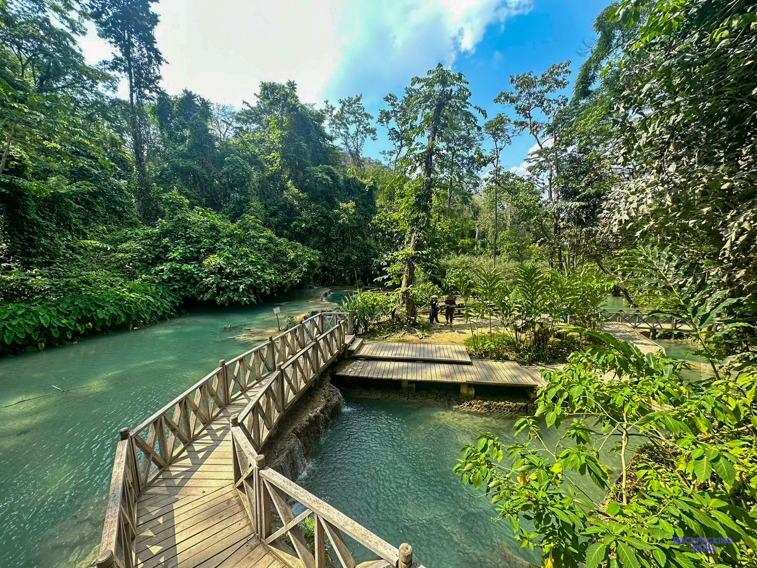 A wooden footbridge crosses the turquoise pools at Kuang Si, surrounded by tall jungle trees.