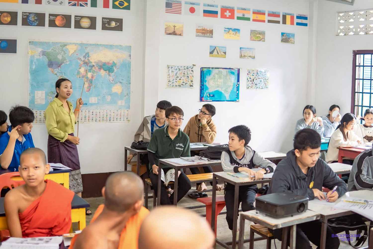 A Lao teacher stands by a world map in front of a class of students and young monks.
