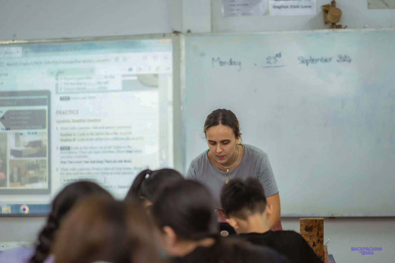 A volunteer helps students in front of a whiteboard showing an English practice exercise.