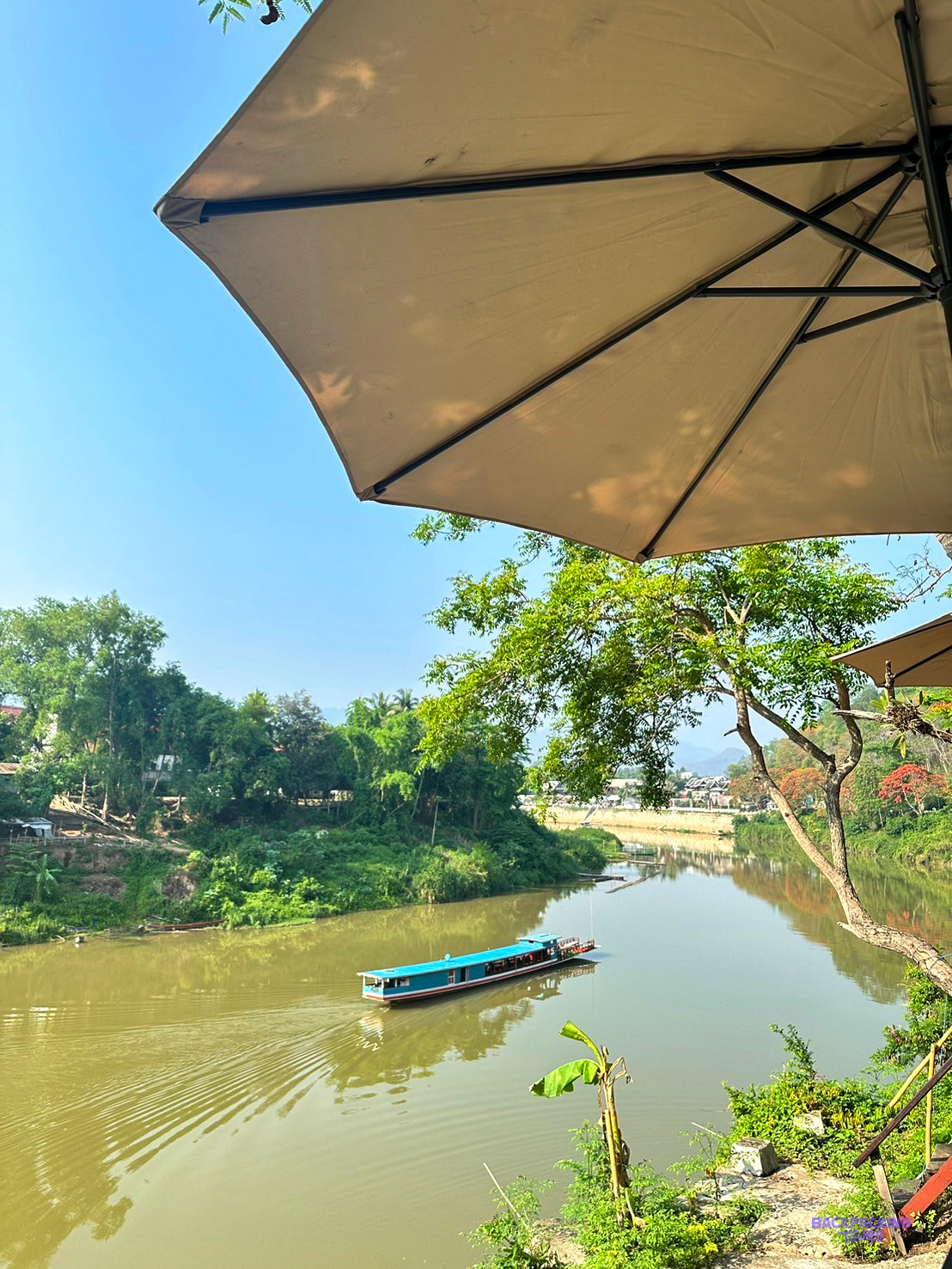 A blue longboat glides down a narrow green river, seen from beneath a riverside café parasol.