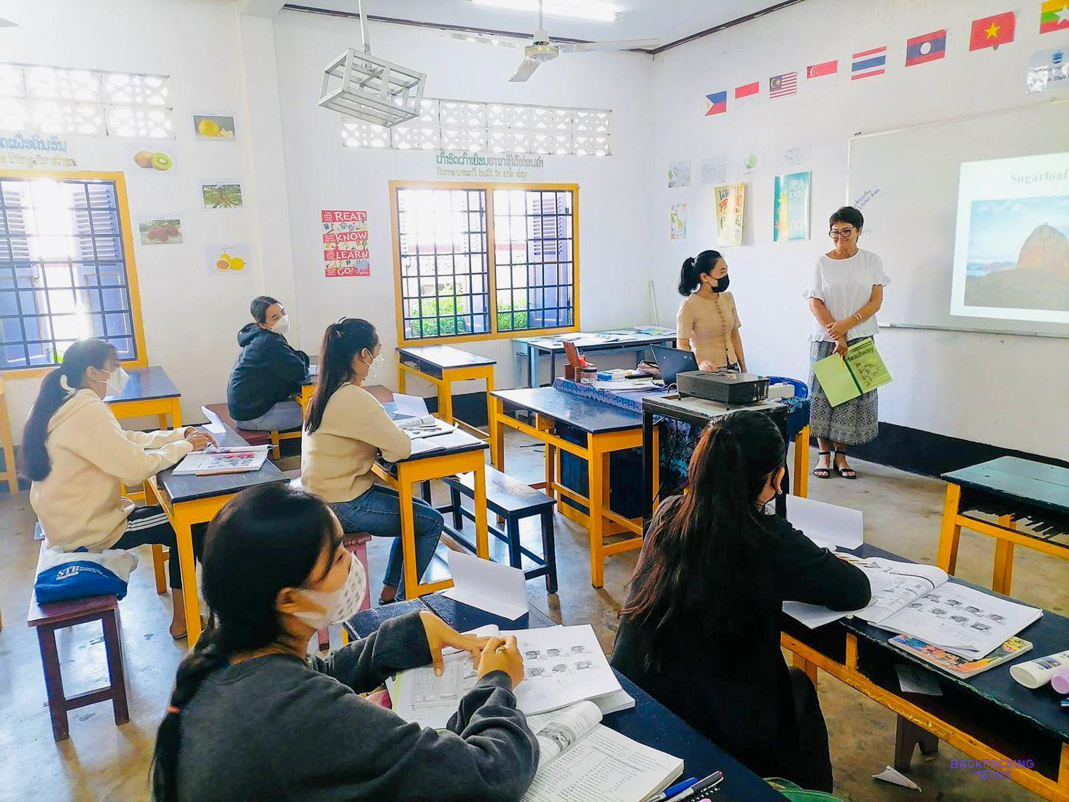 A volunteer and a local teacher stand at the front of a classroom of masked students.