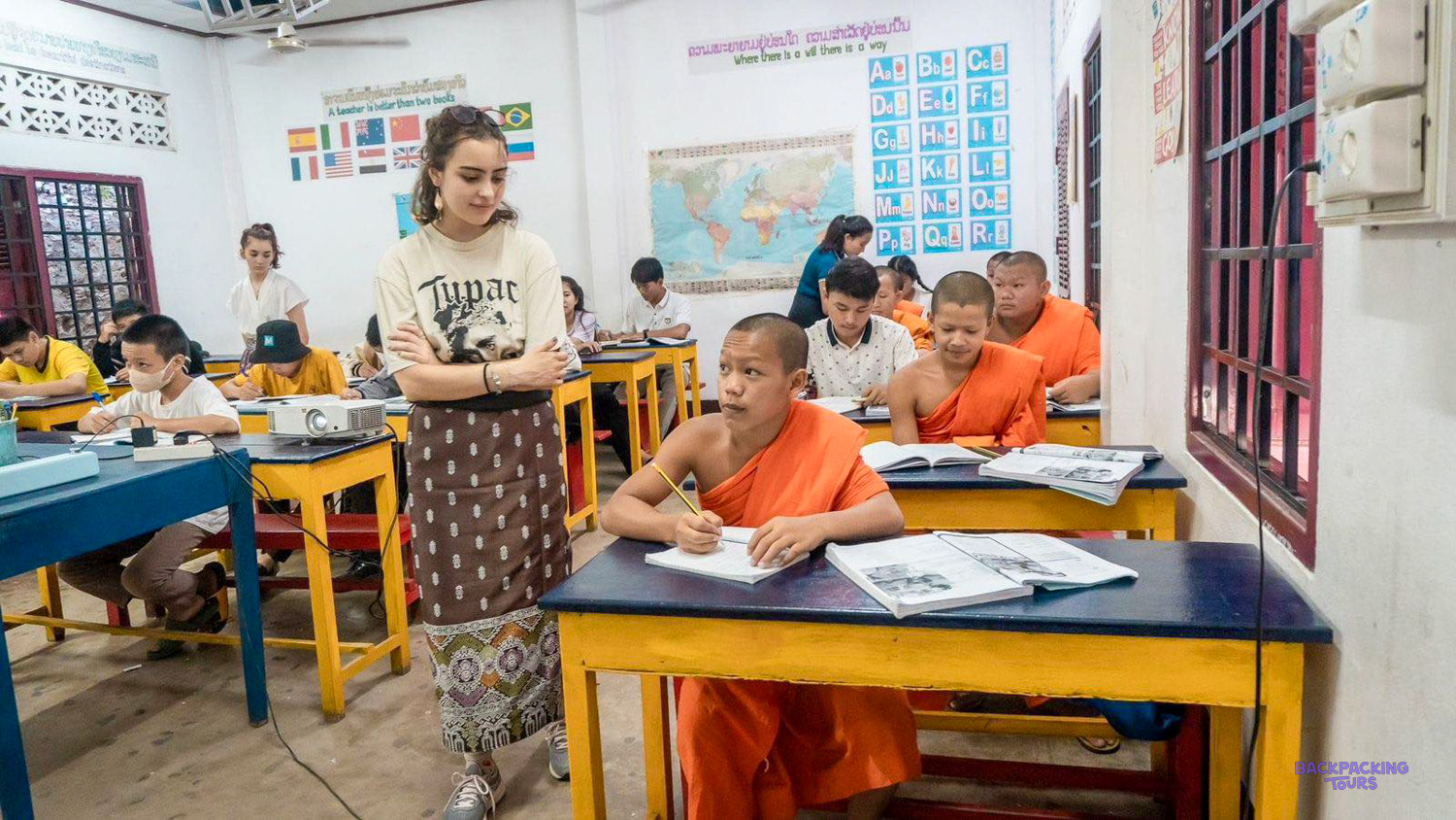 A volunteer stands between desks as novice monks in orange robes work through their English books.