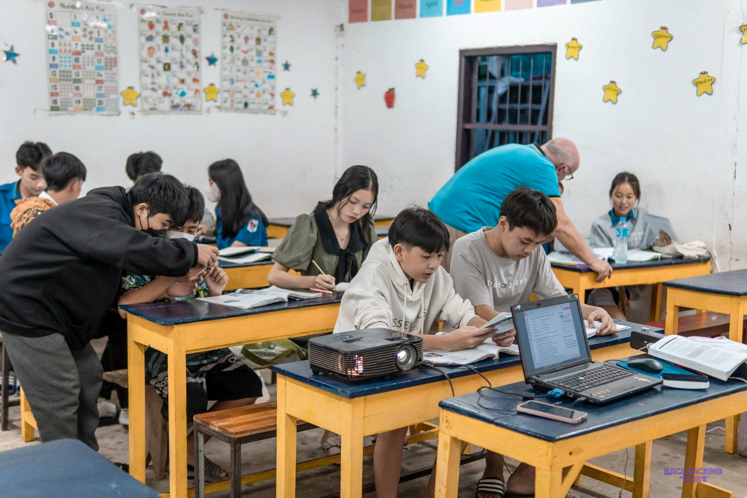 A volunteer leans over to check students' work at yellow desks with a laptop and projector.